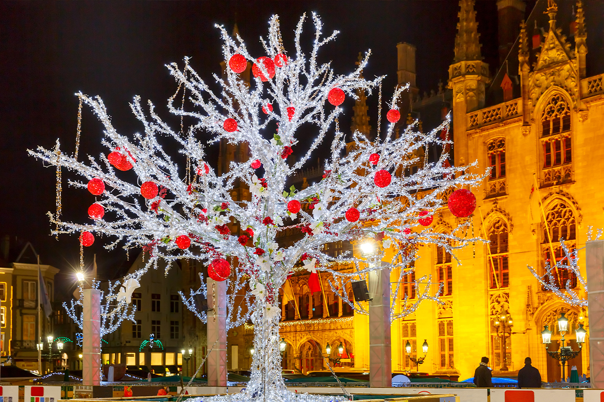 A Christmas tree made of white lights decorated with large red baubles stood in front of buildings